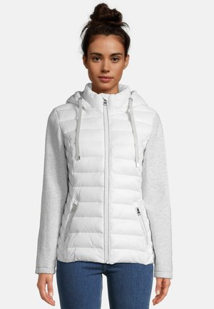 Woman with brown hair in a bun wearing a white quilted jacket with light gray sleeves and blue jeans, facing forward against a plain background.