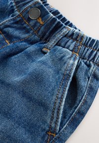 Pantaloni din denim albastru cu talie elastică, având cusături galbene și o mică nasture metalic pe talie, cu buzunare laterale.