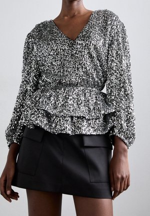 Kvinde iført en langærmet sølv palietter-top med peplum talje kombineret med en sort mininederdel med forlommer.