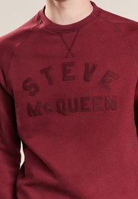 Burgundy sweatshirt with raised embroidered "STEVE McQUEEN" text across the chest, worn by a person shown from the neck to waist.