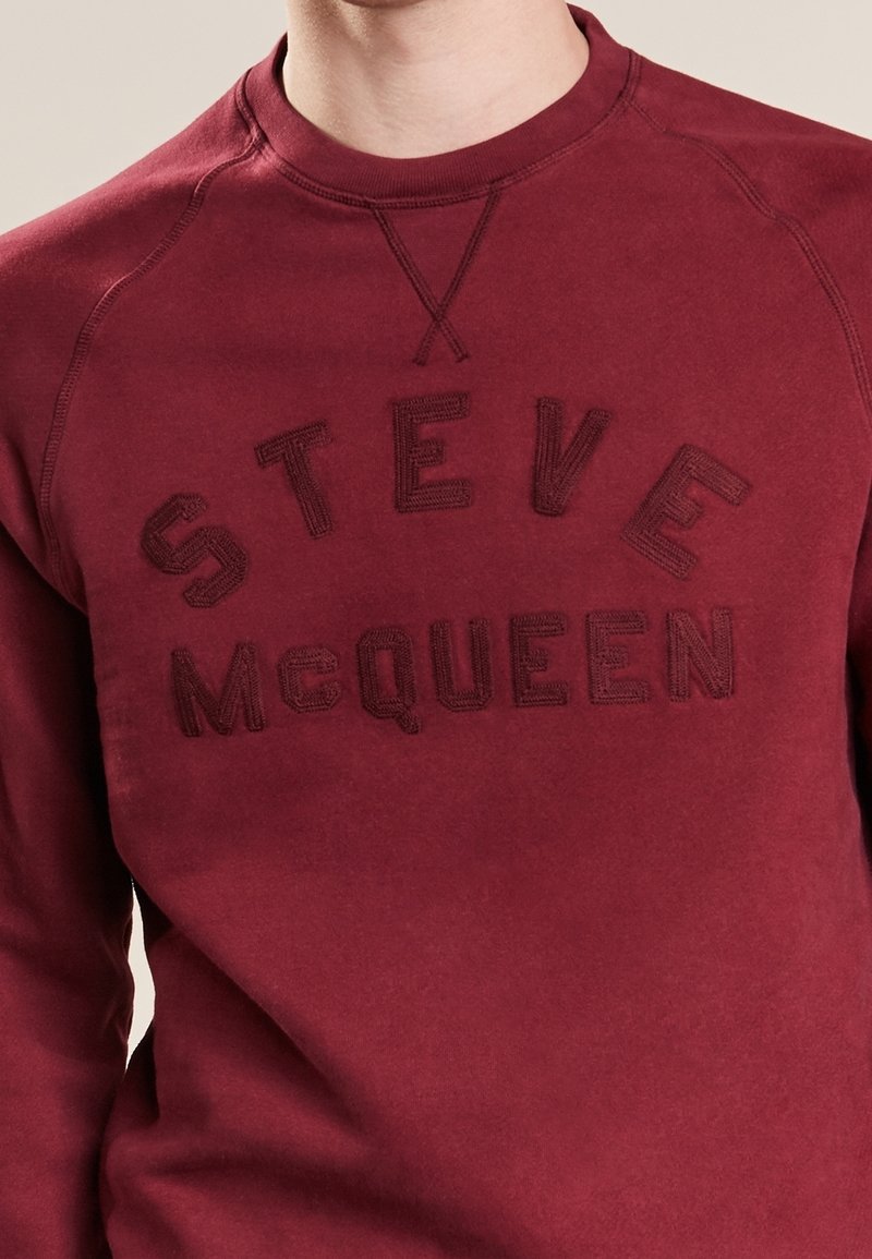 Burgundy sweatshirt with raised embroidered "STEVE McQUEEN" text across the chest, worn by a person shown from the neck to waist.