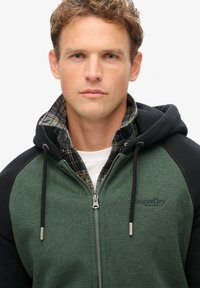 Sudadera con cremallera en verde y negro, de textura suave, que cuenta con una capucha grande, cremallera plateada y un logo en el pecho; colocada sobre una camisa de cuadros.