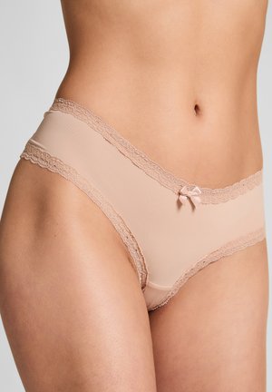 Middendeel van een vrouw die lichtroze ondergoed met kant en een klein strikje op de taille draagt, tegen een effen lichtgrijze achtergrond.