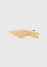 Chaussure à talon haut beige à bout pointu avec bride à la cheville et semelle texturée, vue de dessous sur fond blanc.