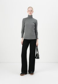 Grijze turtlenecktrui, lange mouwen, gecombineerd met zwarte wijde broek. Model houdt een kleine zwarte handtas vast, staand voor een effen achtergrond.