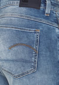 Los vaqueros de mezclilla azul desgastada presentan un bolsillo trasero con un detalle de costura curva y una etiqueta de cuero oscuro en la cintura.