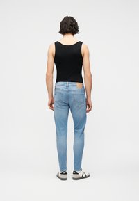 Άτομο με κοντά σκούρα μαλλιά, γυρισμένο πλάτη, φοράει μαύρο αμάνικο τοπ, ανοιχτό μπλε τζιν και άσπρα αθλητικά παπούτσια με μαύρες λεπτομέρειες.