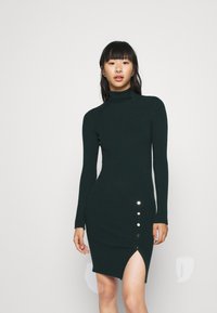 Dunkelgrünes, geripptes Strickkleid mit hohem Kragen, langen Ärmeln und einem seitlichen Schlitz, der drei goldene Knopfakzente hat.