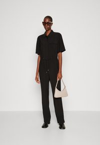 Combinaison noire à manches courtes, avec un col et une ceinture ajustable ; associée à un sac à main beige et des lunettes de soleil rectangulaires marron.
