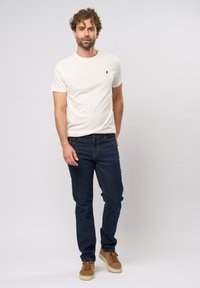 Camiseta de cuello redondo de color beige claro con un pequeño logotipo, jeans oscuros y zapatillas color beige. El modelo está de pie contra un fondo liso.
