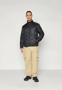 Melna, quiltota virsjaka ar sešstūra rakstu, pilna rāvējslēdzēja, augsts apkakles, un logo uz kreisā krūts. Saskaņota ar bēšām kravas biksēm un melnām kurpēm.