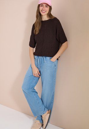 Jeune femme souriante, portant une casquette rose, un pull noir à manches courtes, un jean large bleu clair et des chaussures beige à enfiler, debout contre un mur beige.