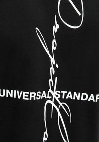Sort stof med stor hvid tekst og kursivde design, herunder "UNIVERSAL STANDARD" og en stiliseret underskriftslignende design.