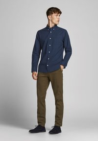 Camisa de botones azul marino con un bolsillo en el pecho, combinada con pantalones chinos verdes olivo y zapatos de ante oscuros. Tejido suave, diseño ajustado.