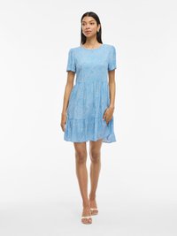 Niebieska sukienka z krótkim rękawem o warstwowym wzorze, z teksturowanym motywem małych białych kropek. Materiał jest lekki i delikatnie opada.