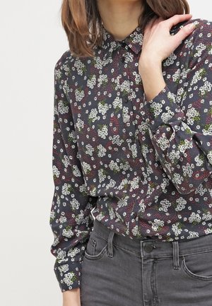 Kobieta w ciemnej, kwiecistej koszuli na długi rękaw zapinanej na guziki, włożonej do wysokich szarych jeansów, z ręką dotykającą szyi.