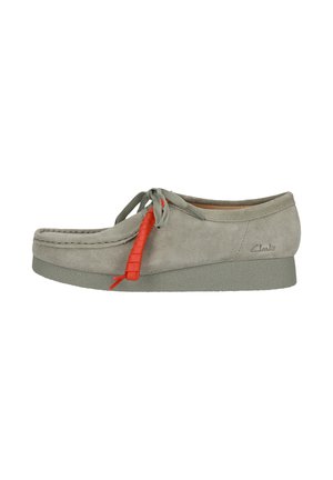 Chaussure en suède gris avec un bout arrondi, dotée d'un lacet rouge contrastant et d'une semelle texturée. La marque est embossée sur le côté.