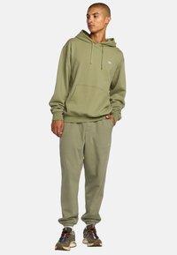 Sudadera verde oliva con cordones y bolsillo tipo canguro, combinada con pantalones de chándal a juego que presentan un pequeño logo y tobillos ajustados.