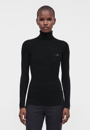Persona con cabello corto que lleva un suéter negro ceñido de cuello alto y pantalones oscuros, de pie frente a un fondo blanco liso.