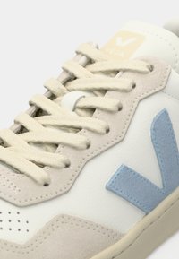 Hvit sneakers med lysegrå semsket skinn-detaljer og en blå semsket logo. Har flate beige snørebånd og perforerte detaljer på tåen.