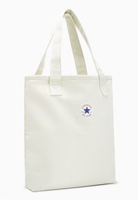 Cream canvas tote bag with two fabric handles and a round logo patch featuring a blue star and red lettering. Square shape, minimal design.