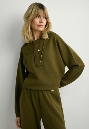 Kobieta o blond włosach nosi oliwkowozieloną bluzę z kapturem z zapięciem na guziki oraz dopasowane spodnie, stoi na tle jasnoszarej ściany.