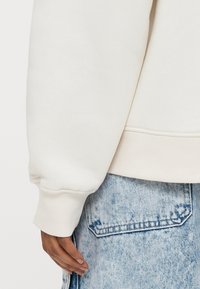 Λευκό μανίκι φούτερ με ριμπ τελείωμα, συνδυασμένο με ανοιχτό μπλε τζιν σορτς που έχει λεπτομέρεια τσέπης. Ορατή απαλή υφή.