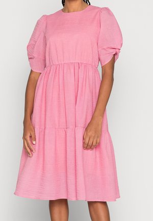 Femme portant une robe rose longueur genou avec des manches courtes bouffantes et une taille froncée, debout sur un fond blanc uni.