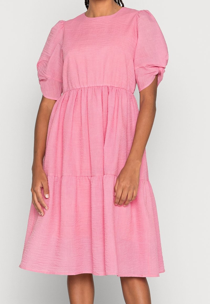 Femme portant une robe rose longueur genou avec des manches courtes bouffantes et une taille froncée, debout sur un fond blanc uni.