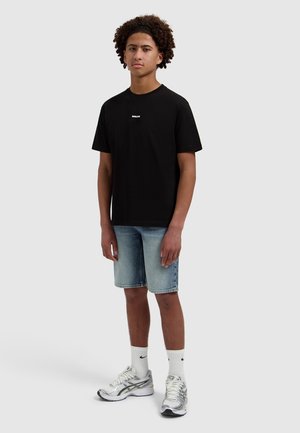 Junger Mann steht, trägt ein schwarzes T-Shirt, hellblaue Jeans-Shorts, weiße Nike-Socken und silberne Laufschuhe.