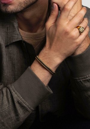 Gold and black bracelet featuring interlocking links, worn on a wrist, paired with a textured gray jacket and beige sweater.