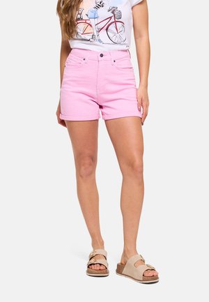 Mujer con pantalones cortos de dobladillo enrollado en rosa claro, camiseta blanca con estampado de bicicleta y sandalias beige con hebilla, de pie frente a fondo blanco.