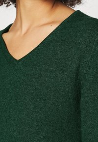 Grüner V-Ausschnitt-Pullover aus strukturiertem Material, das weichen Strick zeigt, mit einer taillierten Passform und einem leicht schimmernden Finish.
