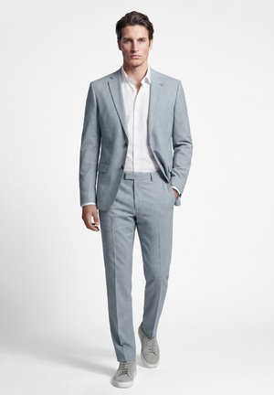 Hombre caminando hacia adelante con traje gris claro, camisa blanca y zapatillas grises a juego sobre un fondo blanco liso.