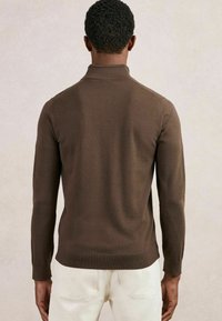 Mann mit kurzen, lockigen Haaren, der einen braunen Pullover mit langen Ärmeln und beige Hosen trägt, steht mit dem Rücken zur Kamera.