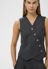 Žena v bezrukávovém tmavě šedém saku na knoflíky a odpovídajících kalhotách, s rukama v kapsách, stojící proti prostému světlému pozadí.