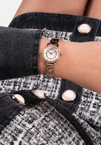 Roosakuldne kell ümmarguse näo, valge diali ja metallise rihmaga. Esitatud randmel, koos tekstuuriga musta ja valge jakiga.