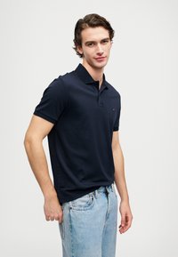 Námořnická polo košile s klasickým límečkem, krátkými rukávy a decentním logem. Spárována se světle modrými džíny, vytvářejícími ležérní outfit.