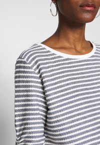 Haut léger à manches longues rayé avec des motifs tricotés alternant gris et blanc, un large col et une texture douce.