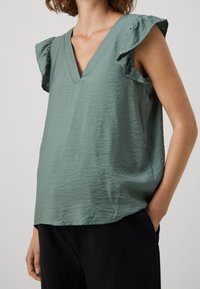 Haut sans manches vert clair avec un col en V, doté d'épaules à volants et d'un tissu doux et texturé. Coupe décontractée avec un ourlet arrondi.