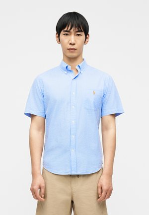 Hombre con camisa de manga corta abotonada a rayas azul claro y blanca, y pantalones cortos caqui, de pie contra un fondo blanco liso.