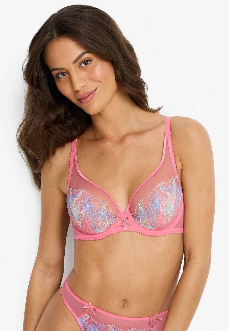 Γυναίκα με κυματιστά καστανά μαλλιά που φορά ροζ ημιδιάφανο σουτιέν και αντιστοιχισμένο εσώρουχο με μπλε κεντήματα λουλουδιών, χαμογελώντας πάνω σε λευκό φόντο.