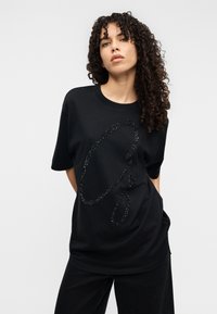 Μαύρο t-shirt με κοντά μανίκια, με μεγάλο, λαμπερό μαύρο διακοσμητικό σχέδιο. Μαλακό ύφασμα με άνετη εφαρμογή, στρογγυλή λαιμόκοψη.