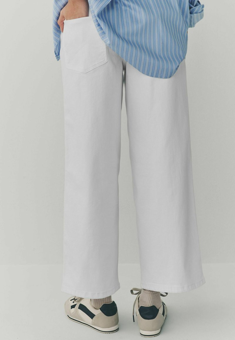 Witte katoenen broek met brede pijpen en een achterzak. Gecombineerd met een blauw gestreept shirt en casual sneakers, met neutrale sokken.