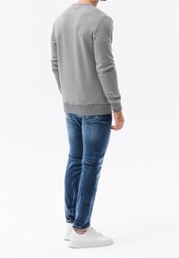 Uomo in felpa grigia, jeans blu e sneakers bianche in piedi con la schiena rivolta verso lo spettatore, su uno sfondo bianco uniforme.