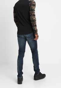 Camisa negra de manga larga con mangas de patrón de camuflaje, combinada con jeans azul oscuro y zapatillas negras, vista desde la espalda.