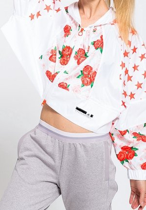 Nainen pukeutuneena valkoiseen lyhytpaitaiseen takkiin, jossa on punaisia ruusu- ja tähtikuvioita, yhdistettynä vaaleanharmaisiin collegehousuihin.