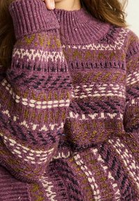 Pull tricoté violet avec des motifs complexes en crème, vert et noir. Présente un col montant côtelé et un tissu épais texturé.