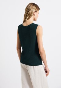 Bez rękawów ciemnozielony sweter z okrągłym dekoltem i teksturowanym materiałem, zestawiony z jasnymi luźnymi spodniami.