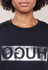 Zbliżenie na osobę z kręconymi włosami, ubraną w czarną koszulę z odwróconym, srebrnym logo "HUGO" na piersi.